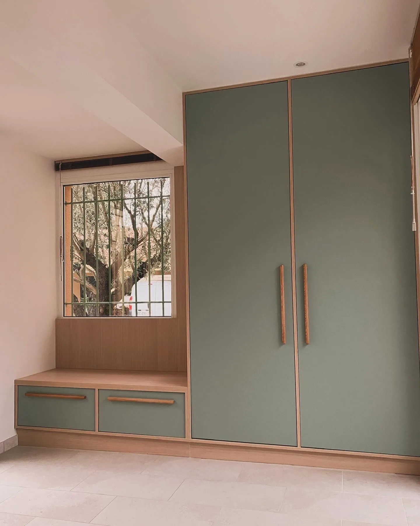 Armoire deux portes vert sauge à chants chêne avec poignées bois, prolongée par un banc-coffre sous la fenêtre.