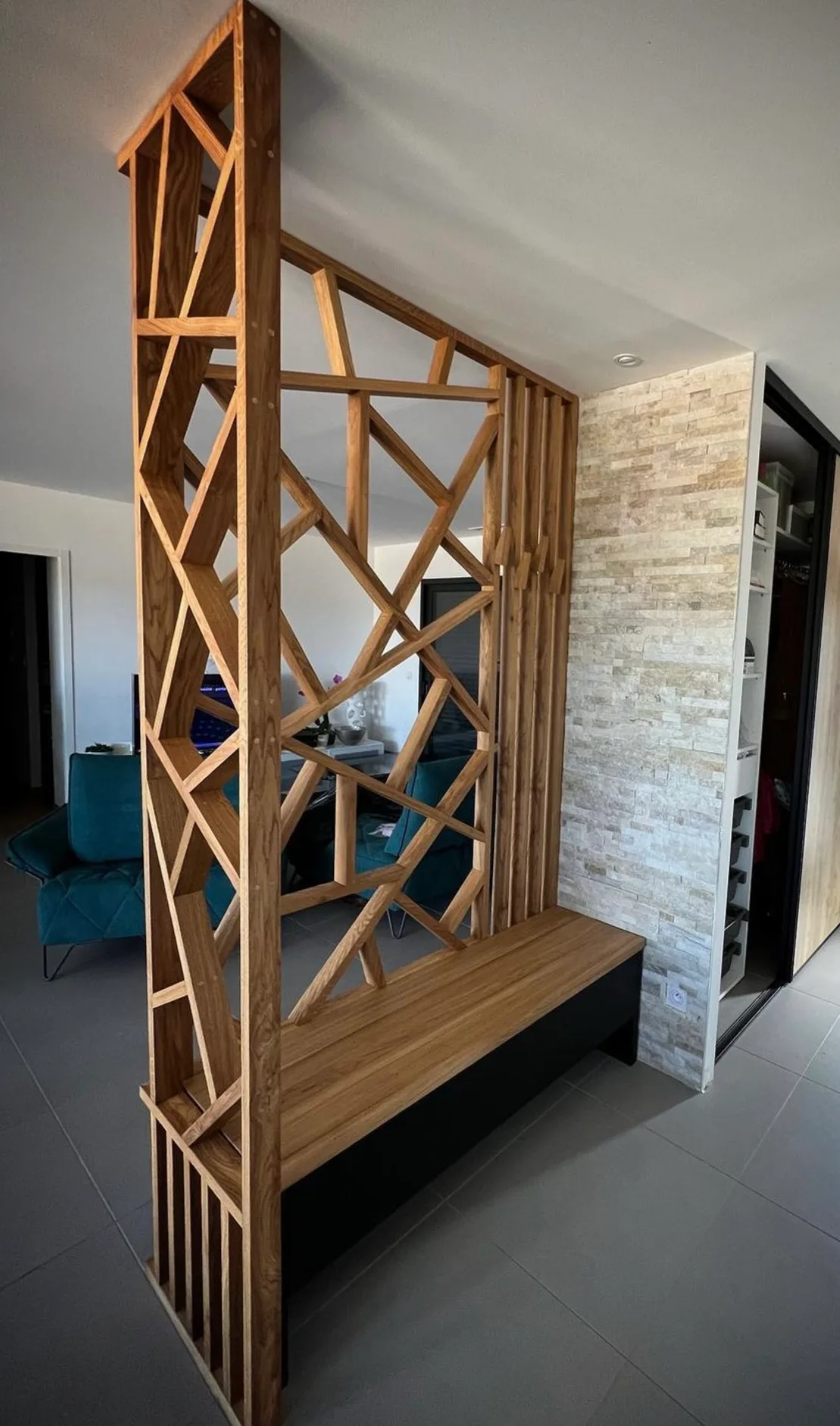 Claustra en chêne à motifs géométriques avec banc d'entrée intégré et socle noir, devant un mur en pierres claires