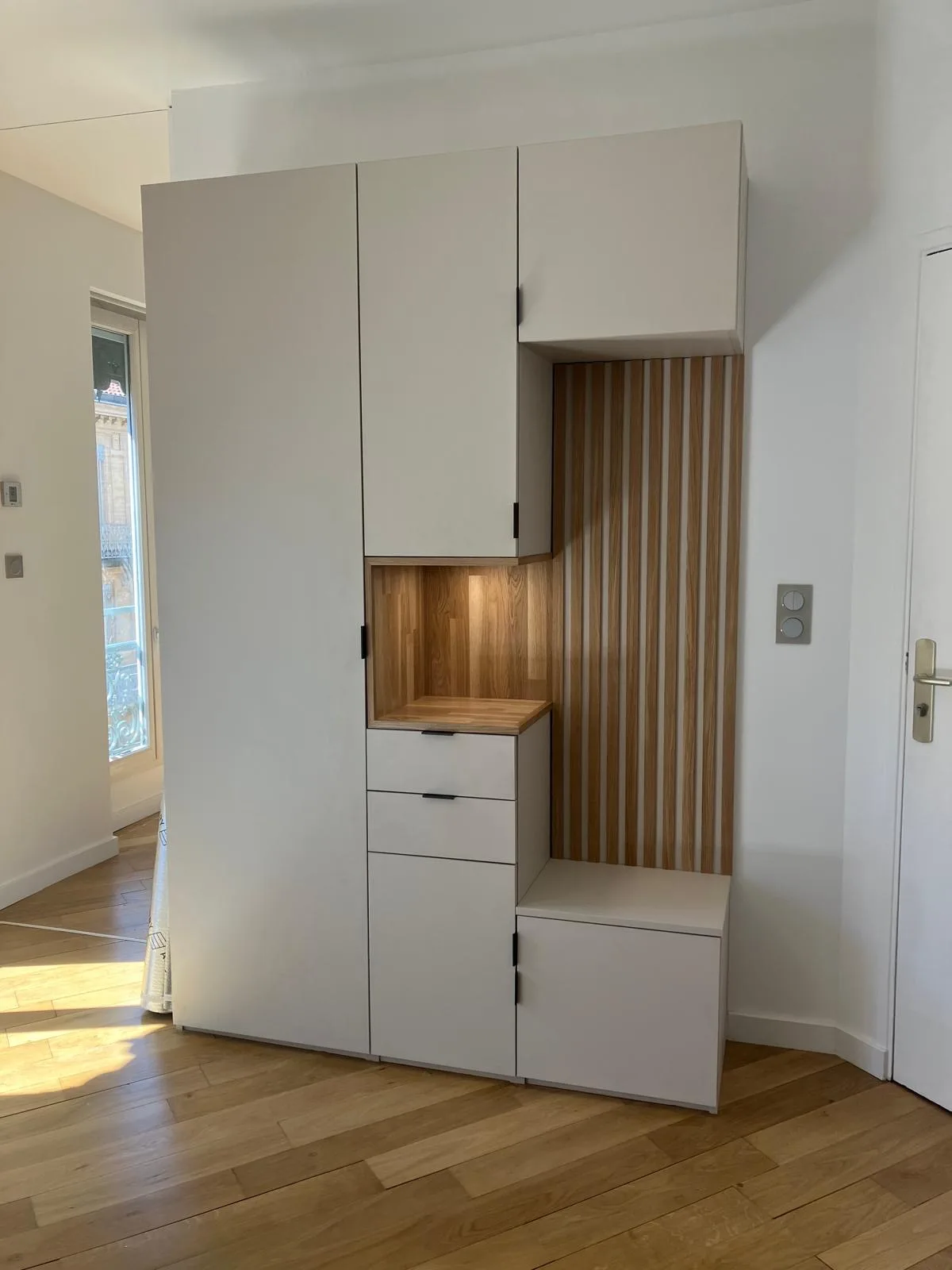 Meuble d'entrée beige toute hauteur avec niche plan en chêne éclairée, tiroirs, banc et claustra en tasseaux