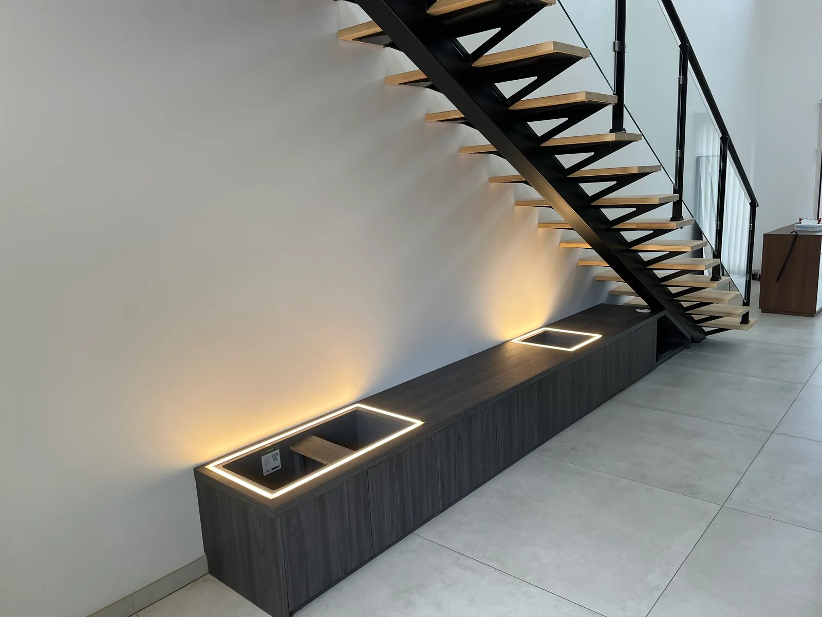 Meuble bas filant gris foncé sous un escalier métallique suspendu, avec niches encastrées éclairées par LED.